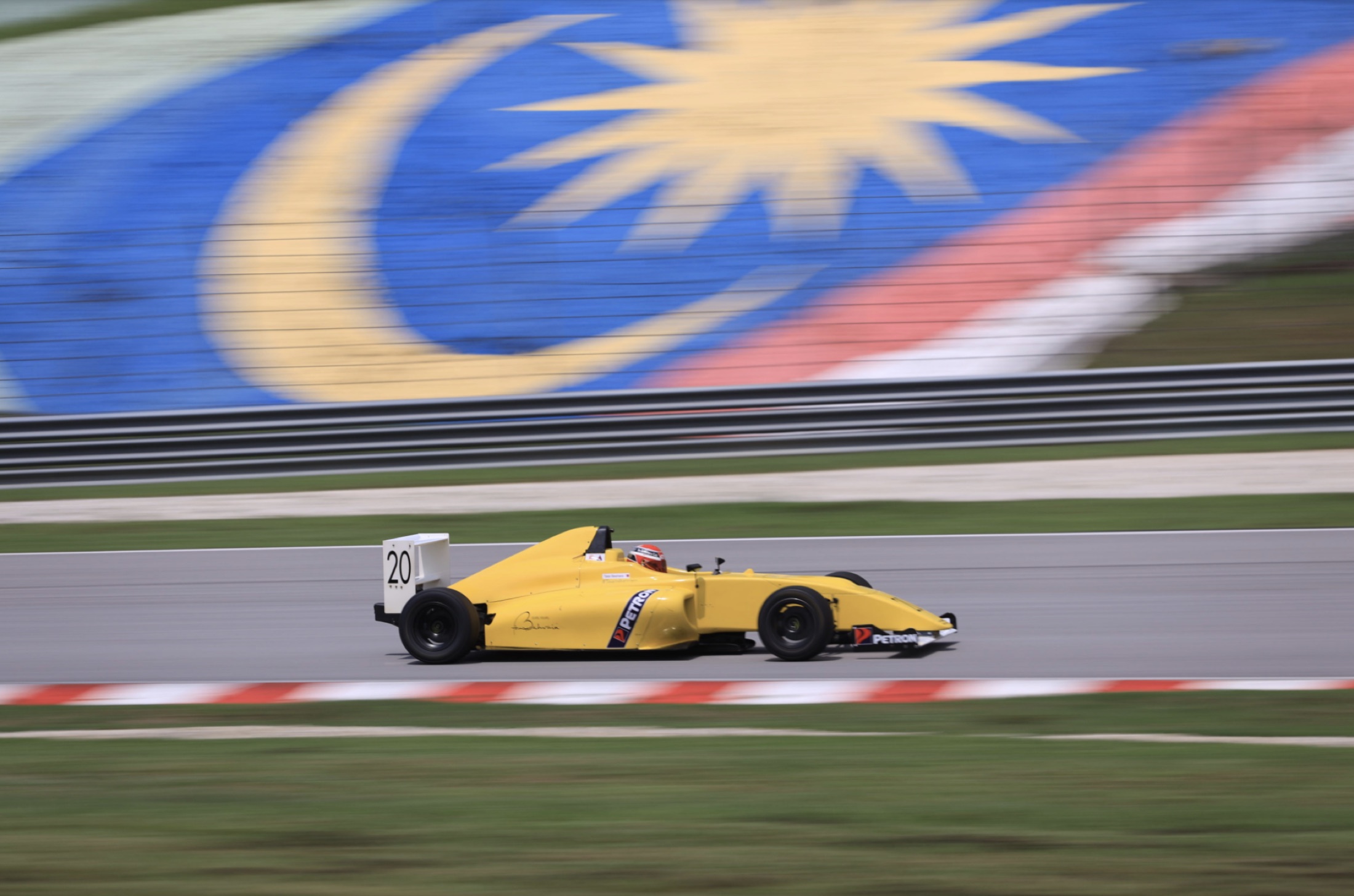 Talal's yellow #20 Formula 4 car passing the Malaysian flag grandstand at Sepang